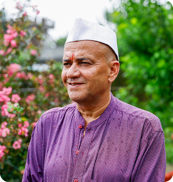 Smiling man in a purple kurta and white cap with green foliage and pink flowers behind him.