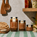 Three jars of PickTown Jackfruit Pickle in different sizes arranged on a green-and-white checkered tablecloth, with wooden utensils, flatbread, and a shelf of pickled vegetables in the background.