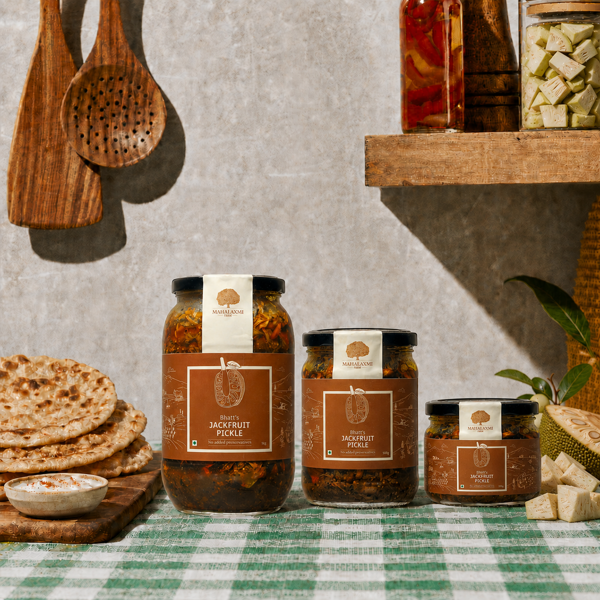 Three jars of PickTown Jackfruit Pickle in different sizes arranged on a green-and-white checkered tablecloth, with wooden utensils, flatbread, and a shelf of pickled vegetables in the background.
