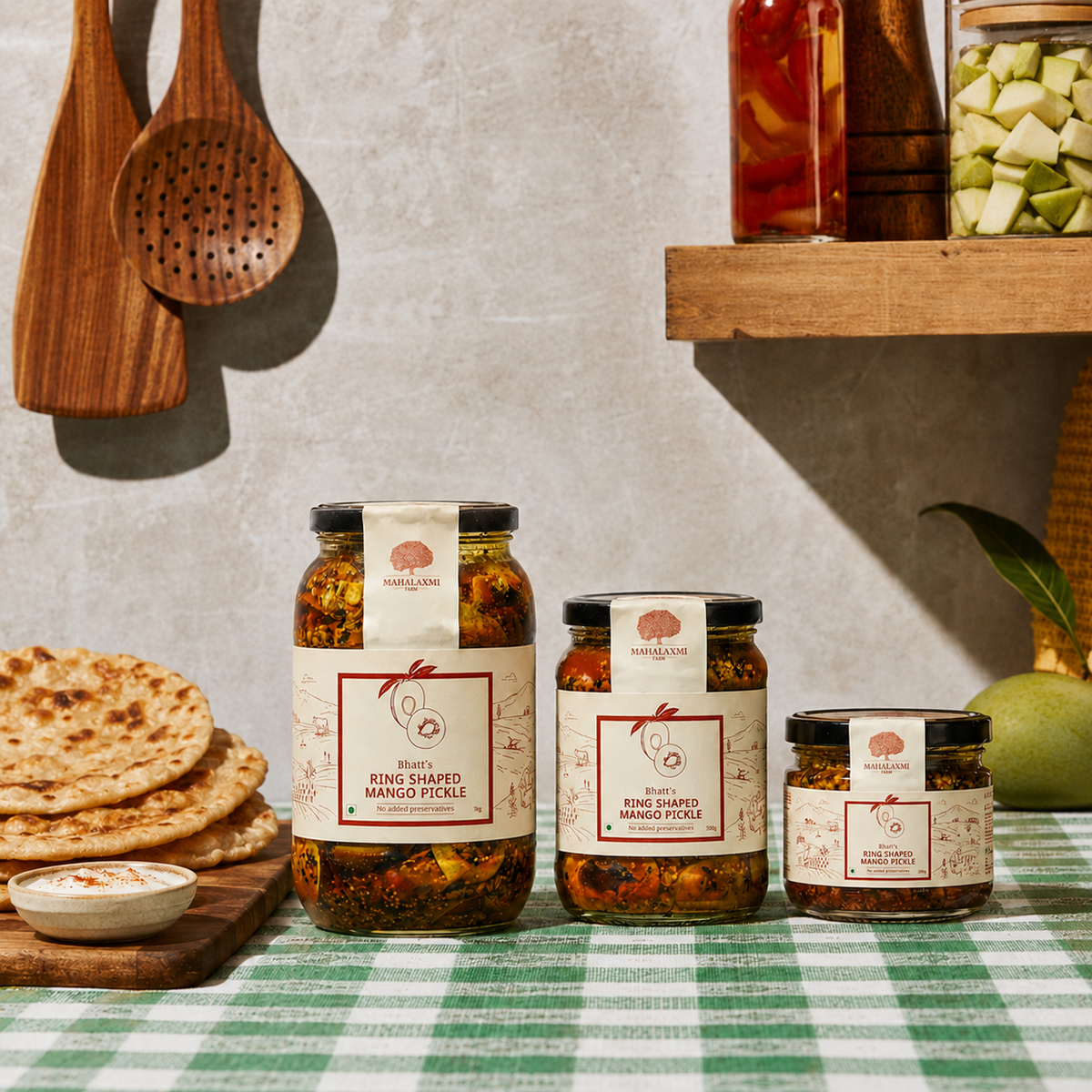 Three sizes of Mahalaxmi Farm Ring Shaped Mango Pickle jars beside flatbread on a green checked tablecloth.