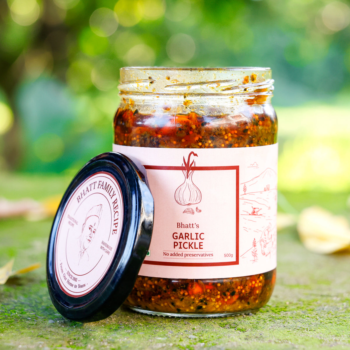 Open jar of Bhatt’s Garlic Pickle with its lid resting beside it against a green garden background.