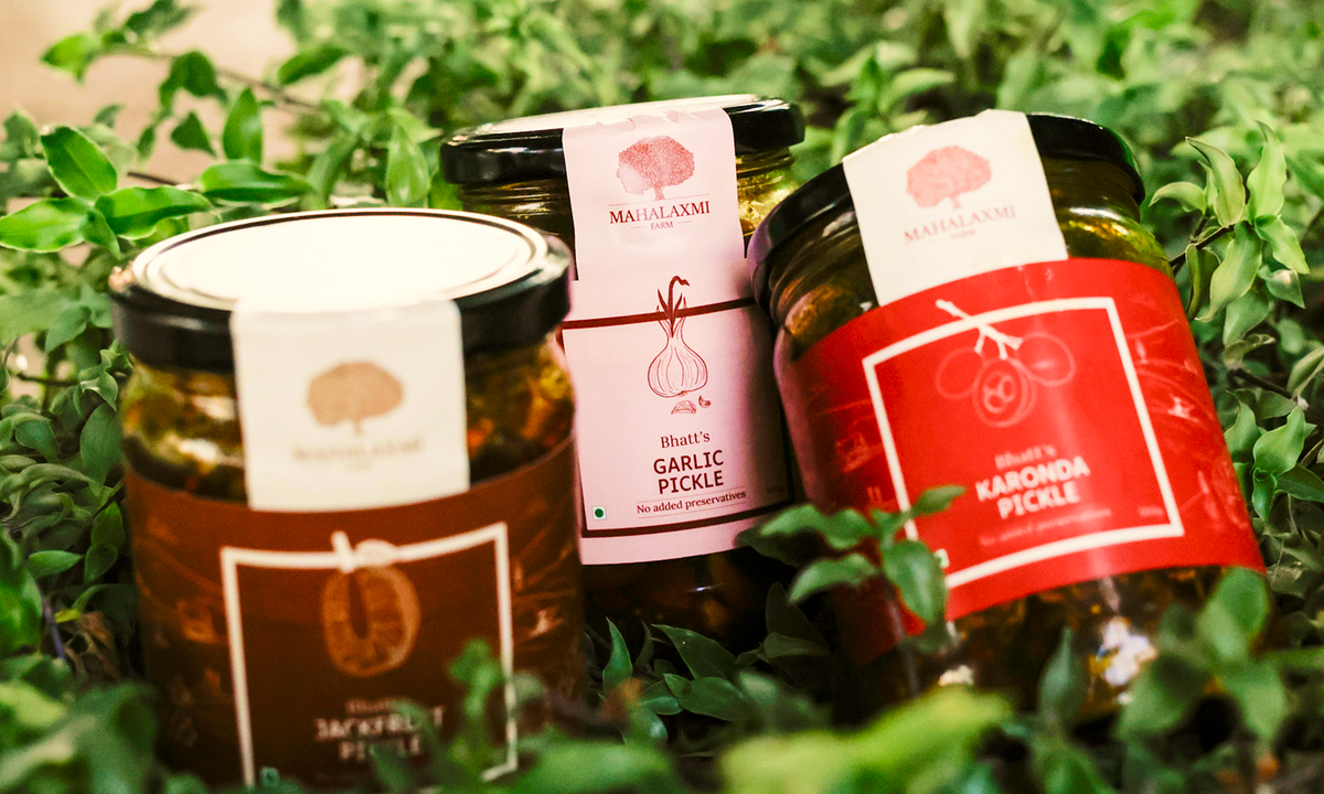 Three glass jars of Mahalaxmi brand pickles nestled in fresh green leaves. The jars include Garlic Pickle with a pink label and Karonda Pickle with a red label, alongside another dark pickle jar with a black lid.