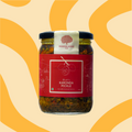 A glass jar of Mahalaxmi Bhatt's Karonda Pickle with a red label and black lid, set against a warm beige background with yellow wavy patterns.