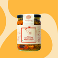 A glass jar of Bhatt's Ring Shaped Mango Pickle by Mahalaxmi, featuring a white label with red text and illustrations, set against a warm yellow and beige abstract background.