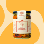 A glass jar of Bhatt's Ring Shaped Mango Pickle by Mahalaxmi, featuring a white label with red text and illustrations, set against a warm yellow and beige abstract background.
