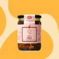 A glass jar of Bhatt's Garlic Pickle by Mahalaxmi Farm, featuring a pink label and a black lid, set against a warm yellow and beige abstract background.