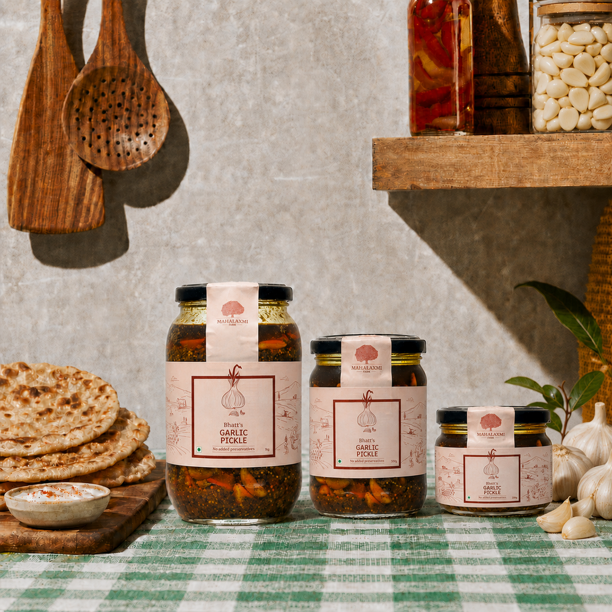Three glass jars of 'The Art of Living' Garlic Pickle arranged on a green and white checkered tablecloth, surrounded by rustic kitchen props including flatbread, garlic, wooden spoons, and a shelf with dried beans.